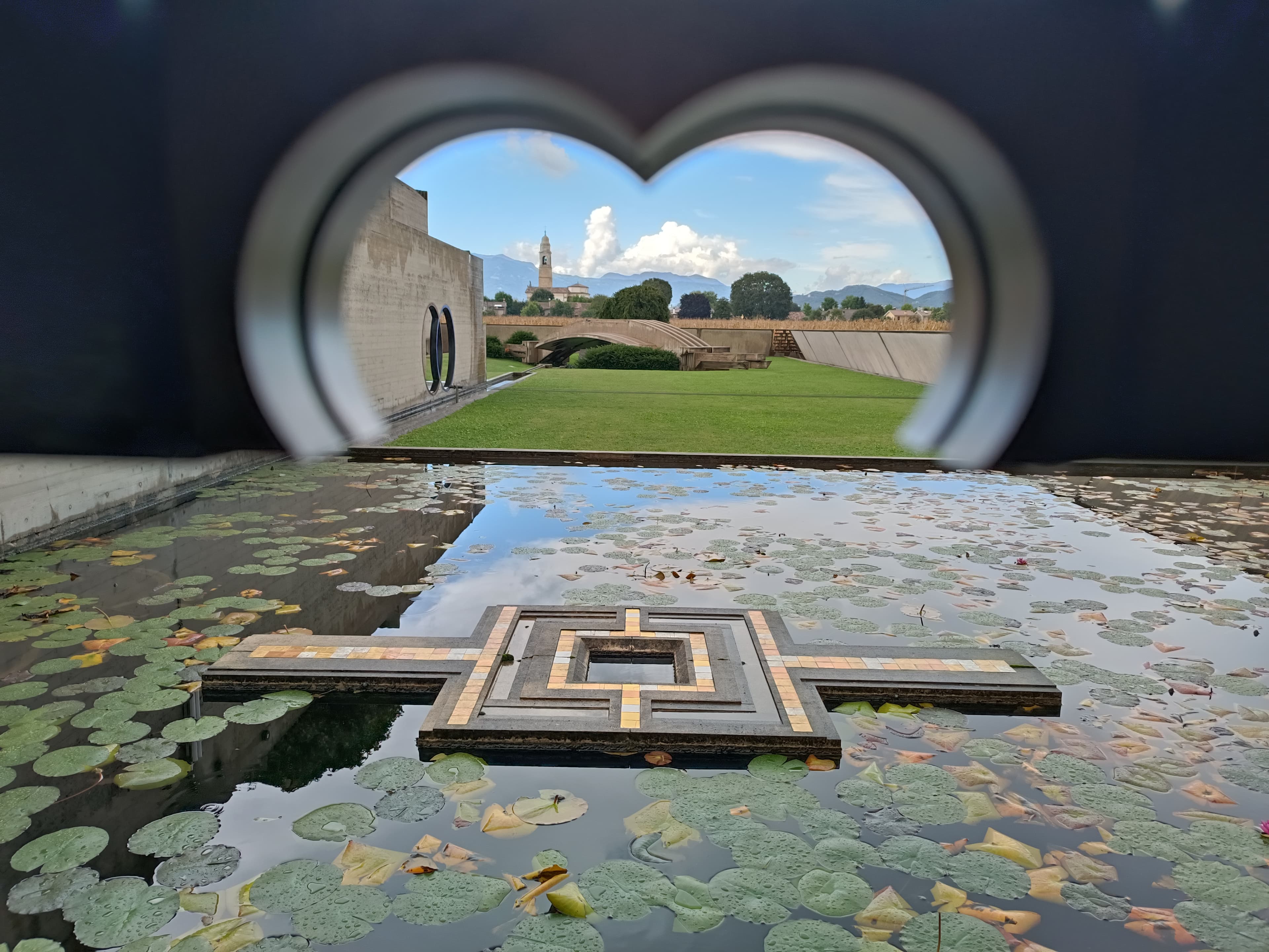 Looking out from inside the Brion Memorial's meditation pavilion through Carlo Scarpa's two interlocking concrete rings, across a lily pond to the cemetery's low boundary wall and the hills beyond.